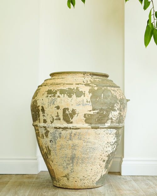 A large rustic ceramic vase with a distressed, peeling cream and gray glaze, standing on a light wood floor against a white wall with green leaves hanging from above.