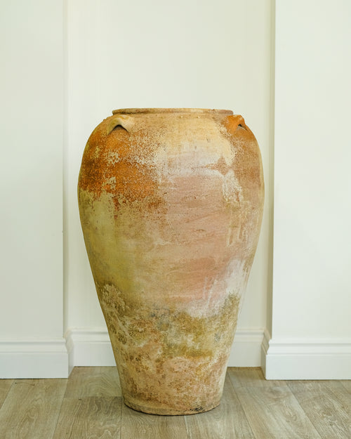 A large rustic ceramic vase with a textured, distressed finish in earthy terracotta, beige, and muted orange tones, standing on a light wood floor against a white paneled wall.