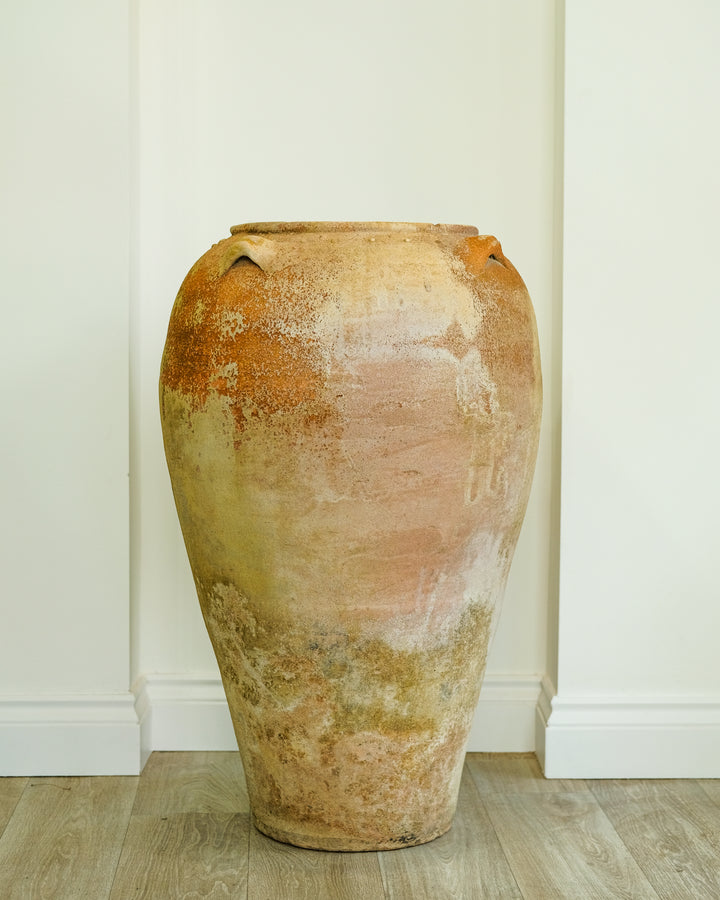 A large rustic ceramic vase with a textured, distressed finish in earthy terracotta, beige, and muted orange tones, standing on a light wood floor against a white paneled wall.