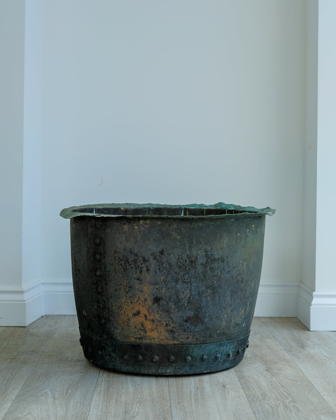 A large vintage cast iron planter pot with a rustic, weathered patina, featuring a green-tinted rim and a band of rivets near the base, set on a light wood floor against a white wall.
