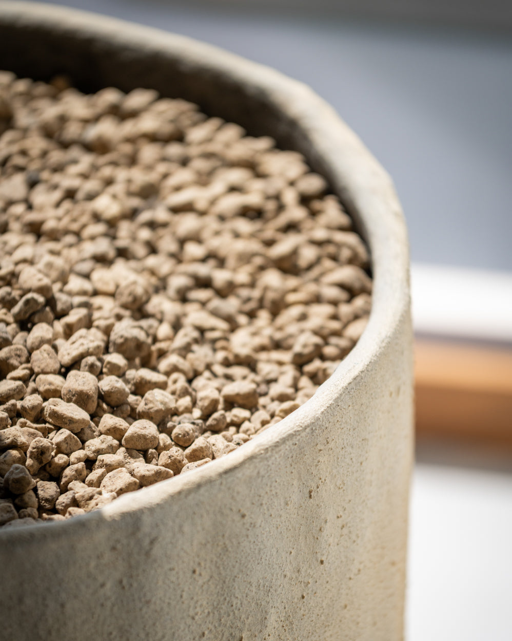 Close-up of a cylindrical concrete planter filled with small tan pebbles, photographed in soft natural light with a blurred neutral background.
